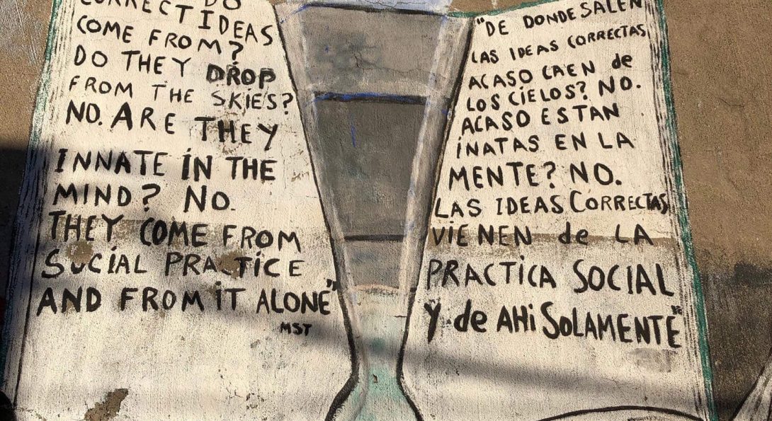 A painting of an open book has an upside down triangular object in the middle. Text written in English and Spanish include sentences such as “Where do correct ideas come from?” amd “De donde salen las ideas correctas acaso caen de los cielos?