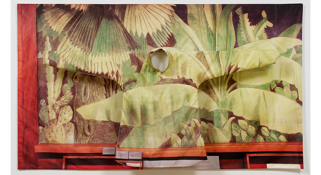 A photograph of a textile against a thick wooden frame. The textile is a drawing of various green plants. Placed over top of the textile is a long sleeve garment in the same pattern.
