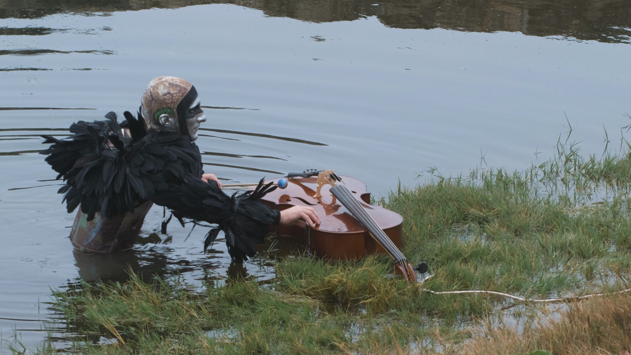 A figure wearing feathers and a helmet stands partly submerged in water and plays a cello that floats in the same water.||A medium-skinned person with long dark hair looks just to the side of the camera.