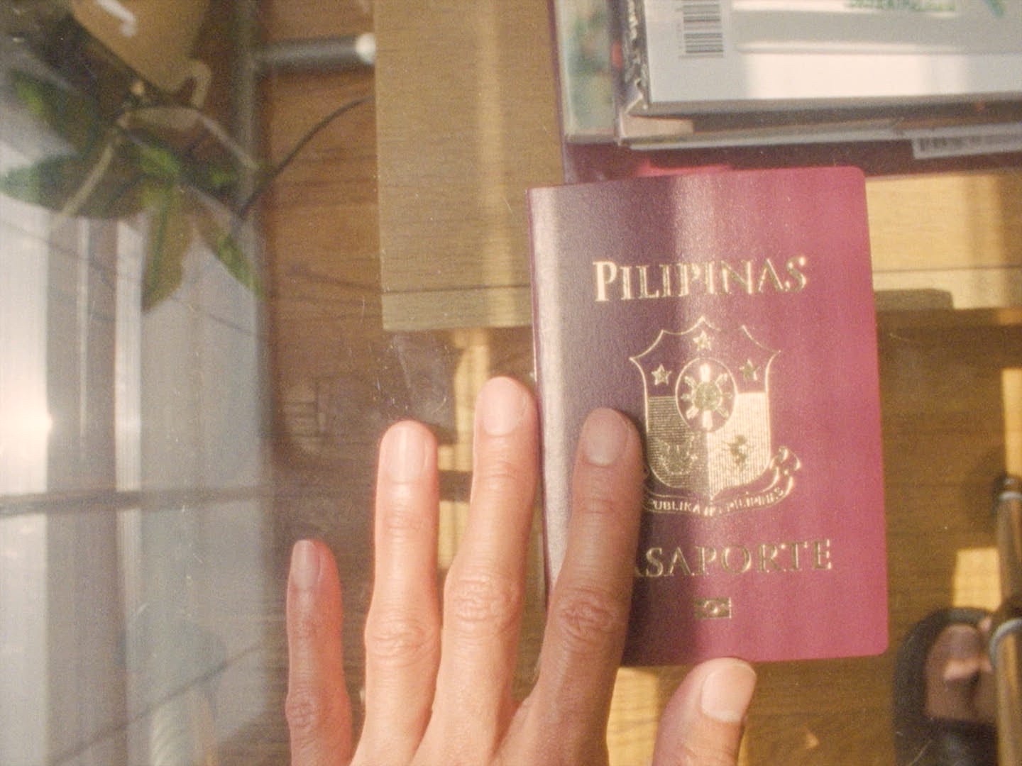 Image of a brown hand touching a burgundy passport with gold font that reads “Pilipinas Pasaporte” and features the Filipino crest in the center. The passport is sitting on a glass table- beneath it there are a stack of books sitting on wood floor.|